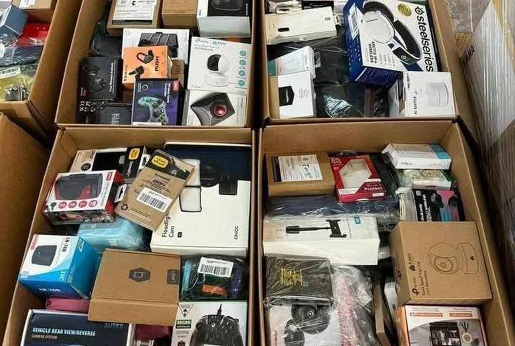Quatro caixas de papelão abertas no chão de um armazém, cheias de pequenos eletrônicos e acessórios